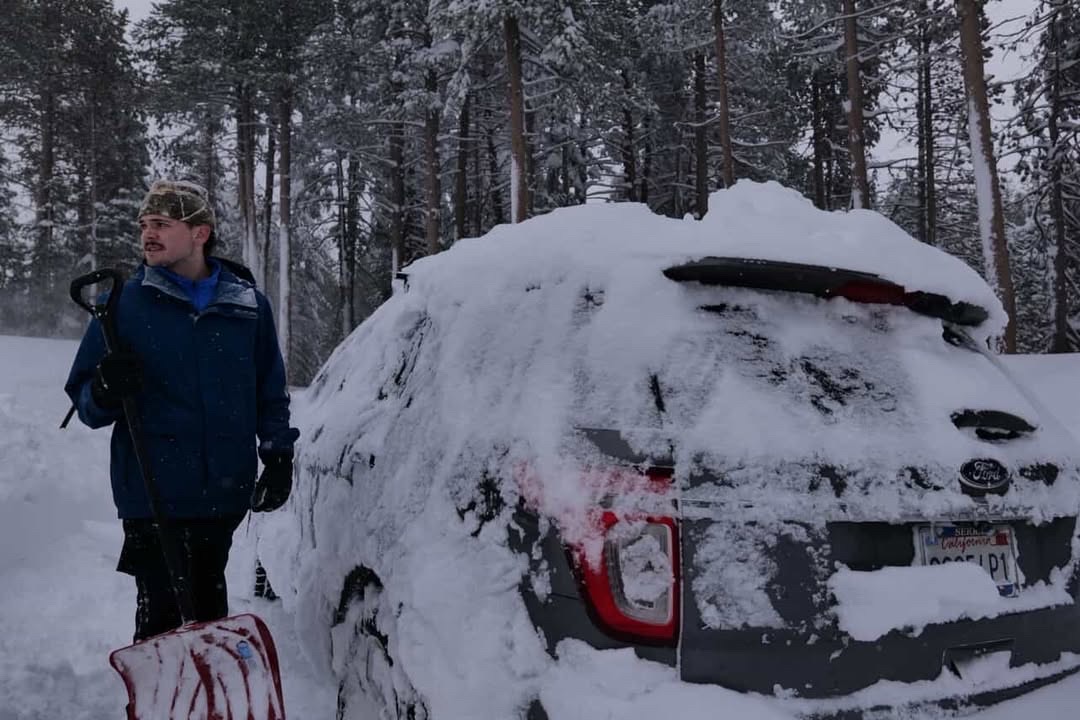 Colin McCarthy holding a snow shovel beside a snow-covered vehicle in a forest